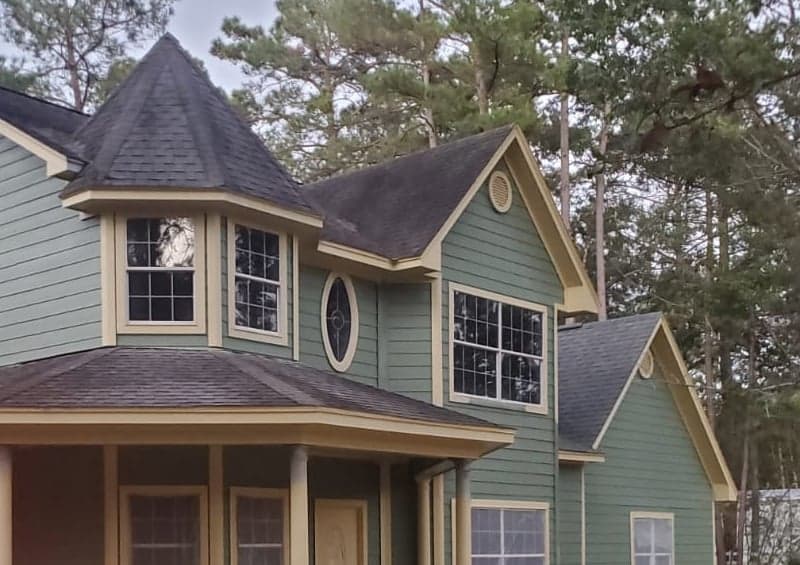 Two-story Magnolia home with green lap siding, yellow trim, a turret roof and a wraparound porch among pine trees