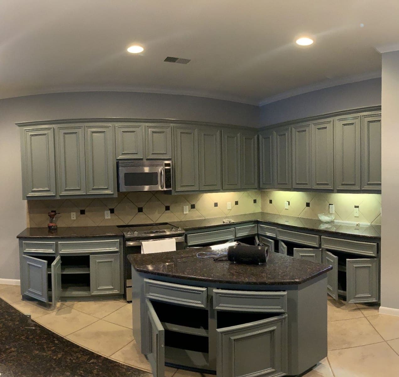 Wide view of the sage green kitchen cabinets and island, doors open while the finish dries