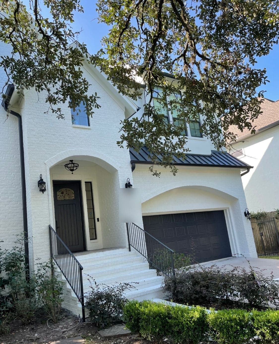 Two-story West University home with white painted brick, an arched entry and a dark garage door under oak branches